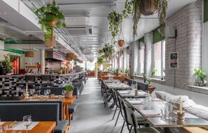 Un restaurant avec de hauts plafonds, des plantes suspendues, des murs en briques et différents types de sièges, notamment des tables et des banquettes.
