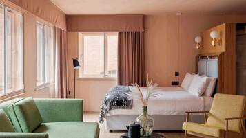 Habitación de hotel con una cama, un sofá verde, un sillón amarillo, una mesita de noche con un jarrón y una ventana con cortinas.