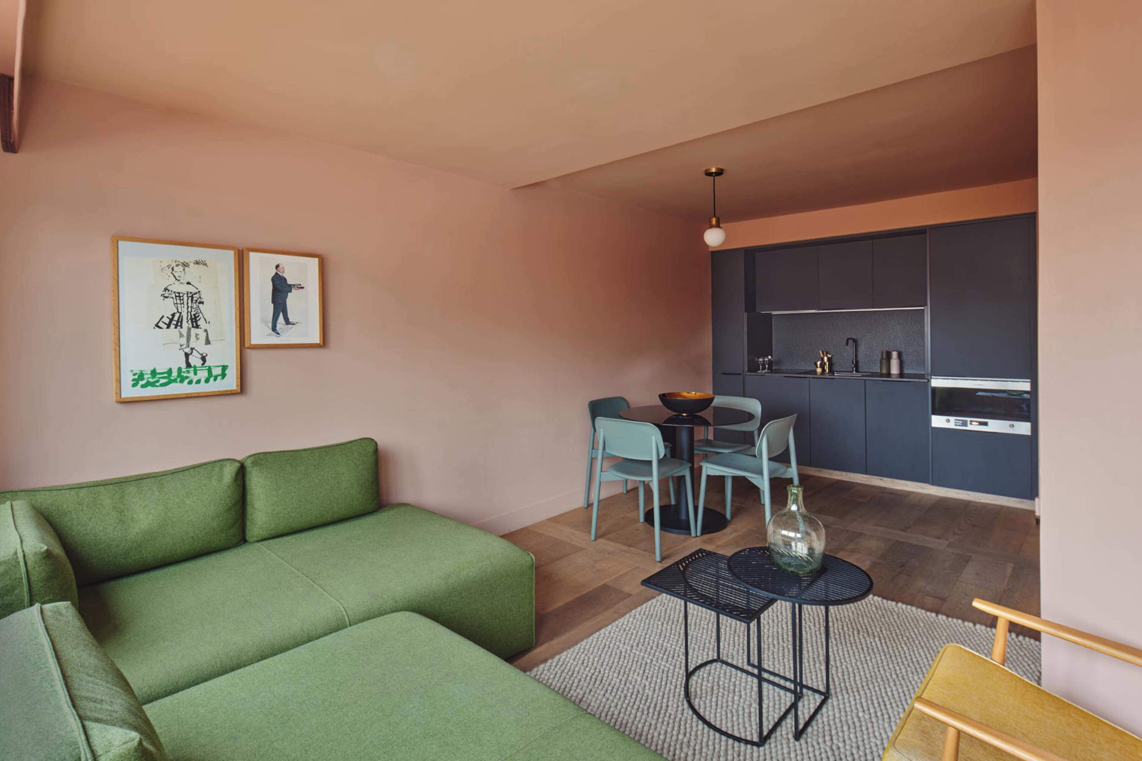 Een moderne woonkamer met een groene bank, een eethoek met een ronde tafel en stoelen, en een keuken met donkere kasten en apparatuur.