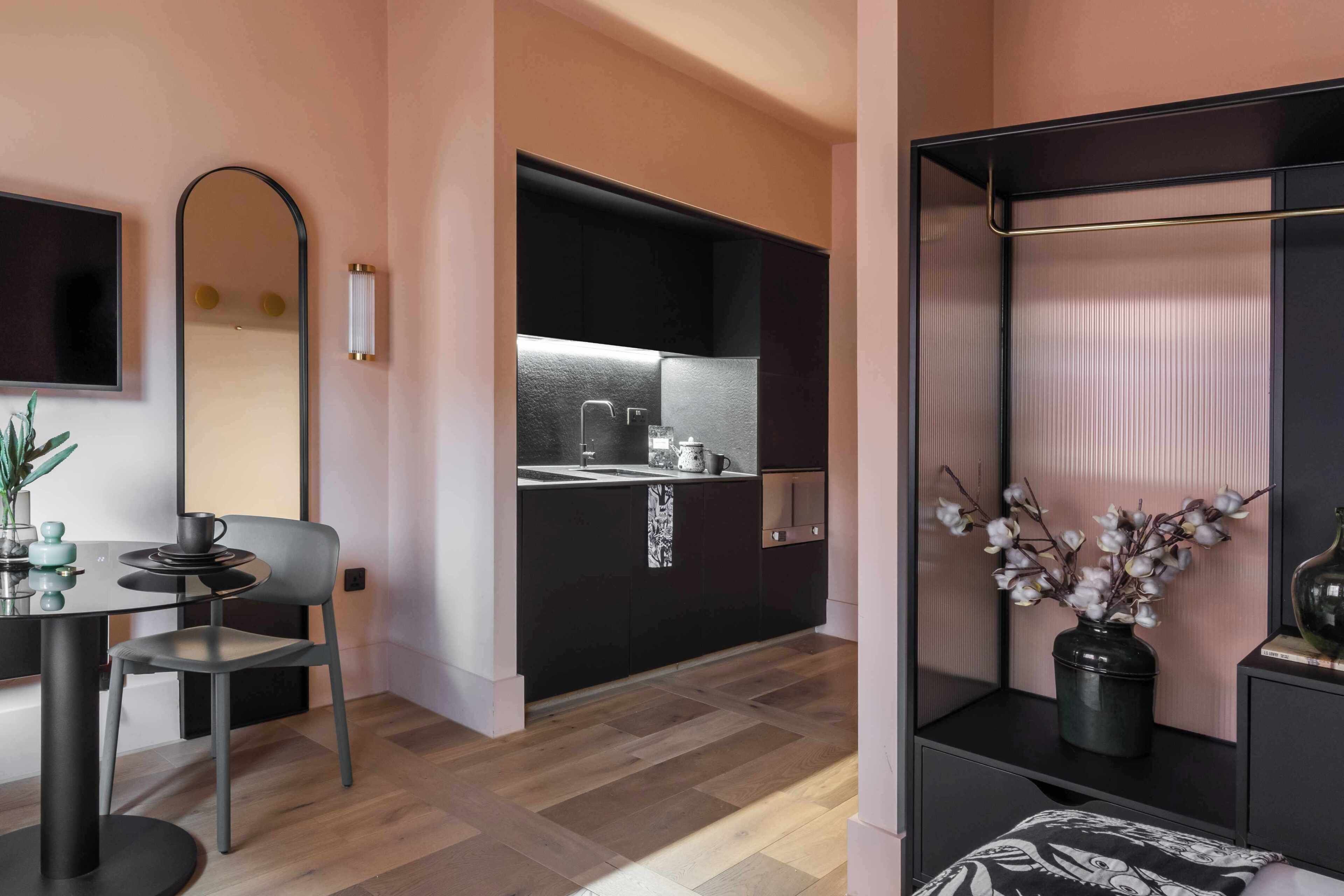 A modern room with a dining table, a chair, a mirror, a black-framed TV, a kitchen area with a sink and cabinets, and a shelf with a vase and decorative items.