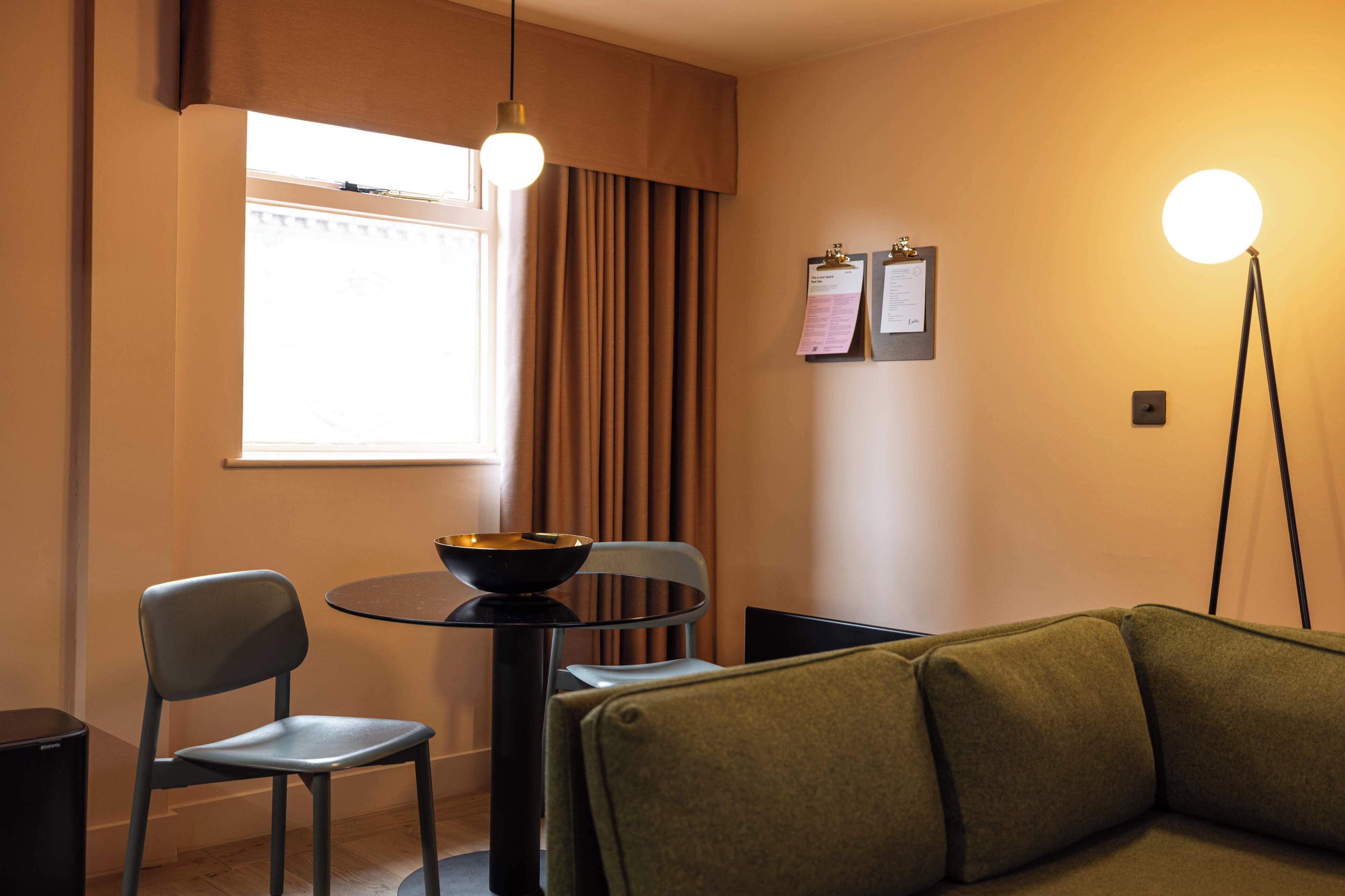 Ein Zimmer mit einem runden Tisch, zwei Stühlen, einem Sofa, einer Schüssel auf dem Tisch, einer Lampe, einem Fenster mit Vorhängen und zwei Pinwänden mit Papieren.