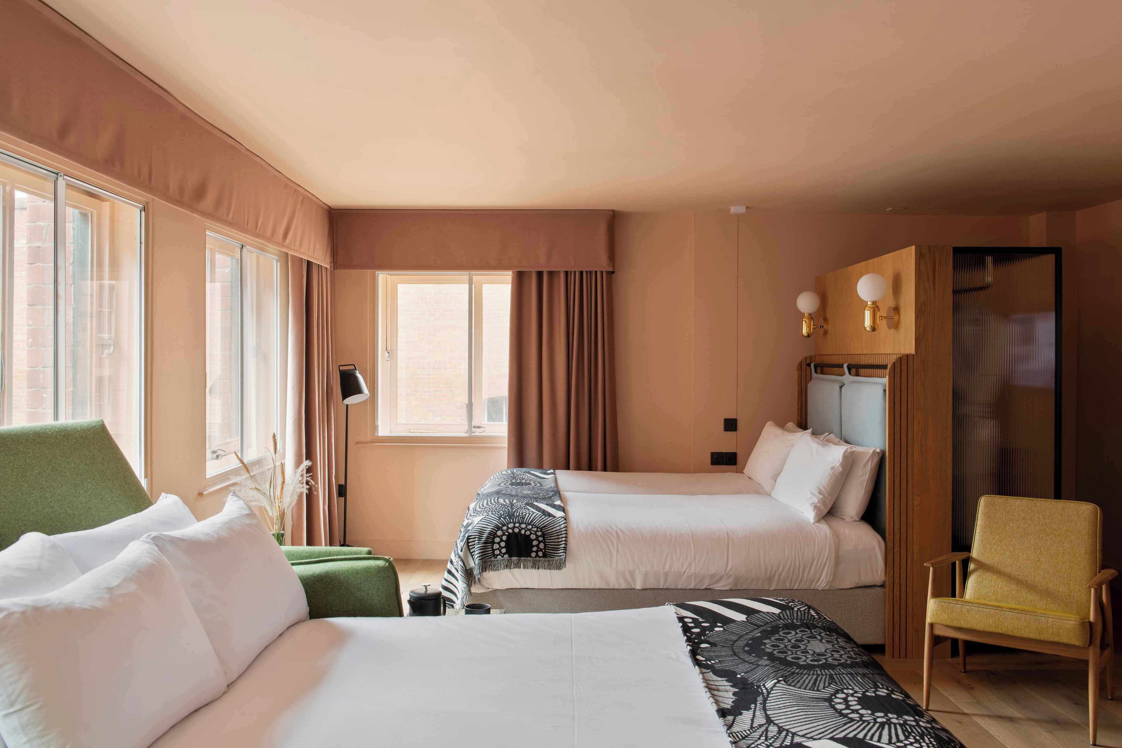 A hotel room with two beds, a green armchair, a yellow armchair, a window with curtains, and a wall-mounted lamp.