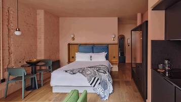 Quarto com cama, mesas de cabeceira, uma cadeira, uma mesinha e um espaço com cozinha compacta.