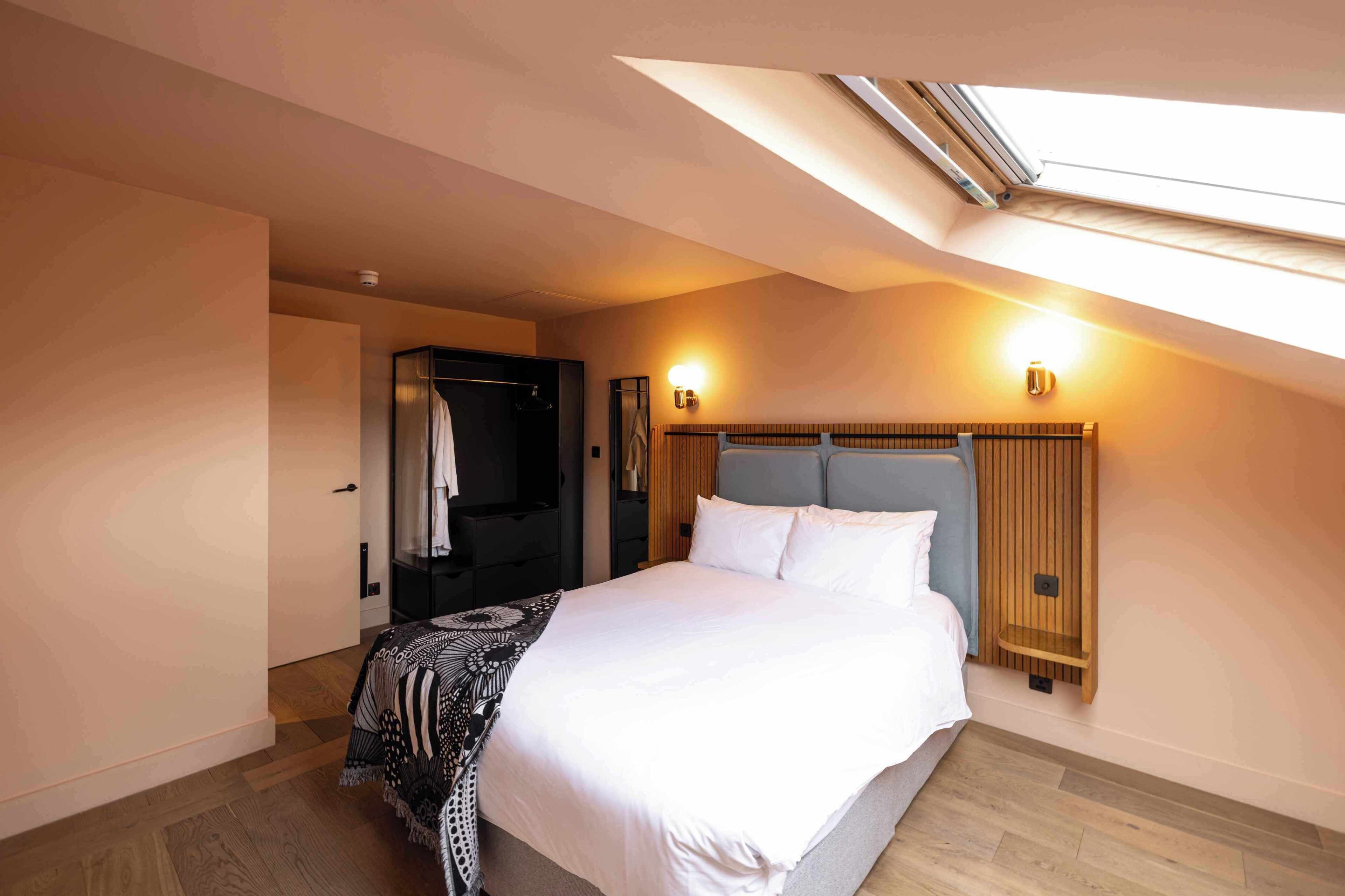 Een slaapkamer met dakraam, een bed met witte beddengoed, een houten hoofdeinde en een nachtkastje.