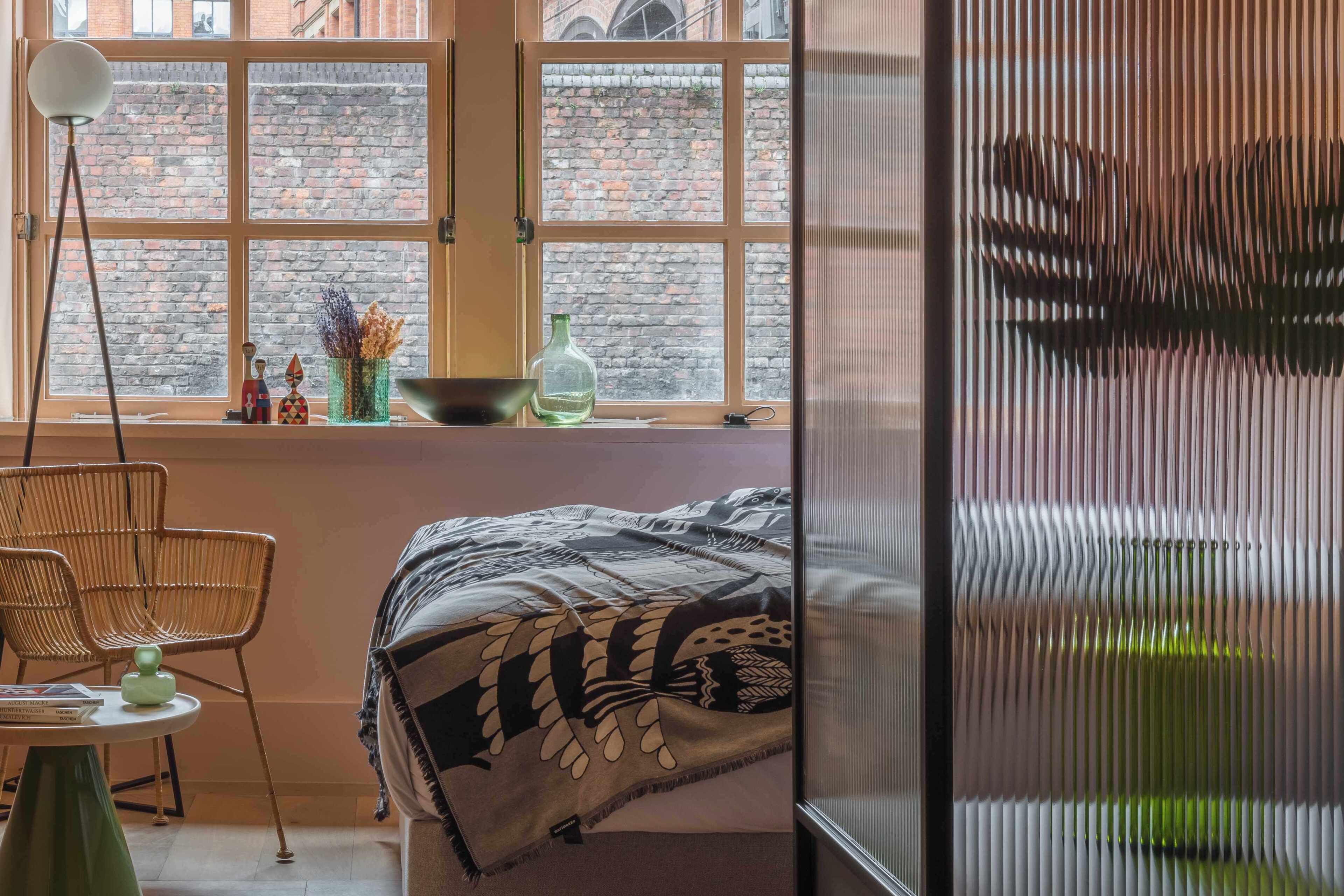 Ein Zimmer mit einem Bett, einem Fenster mit Blick auf eine Ziegelwand, einem Sessel und einem Tisch mit einer Lampe.