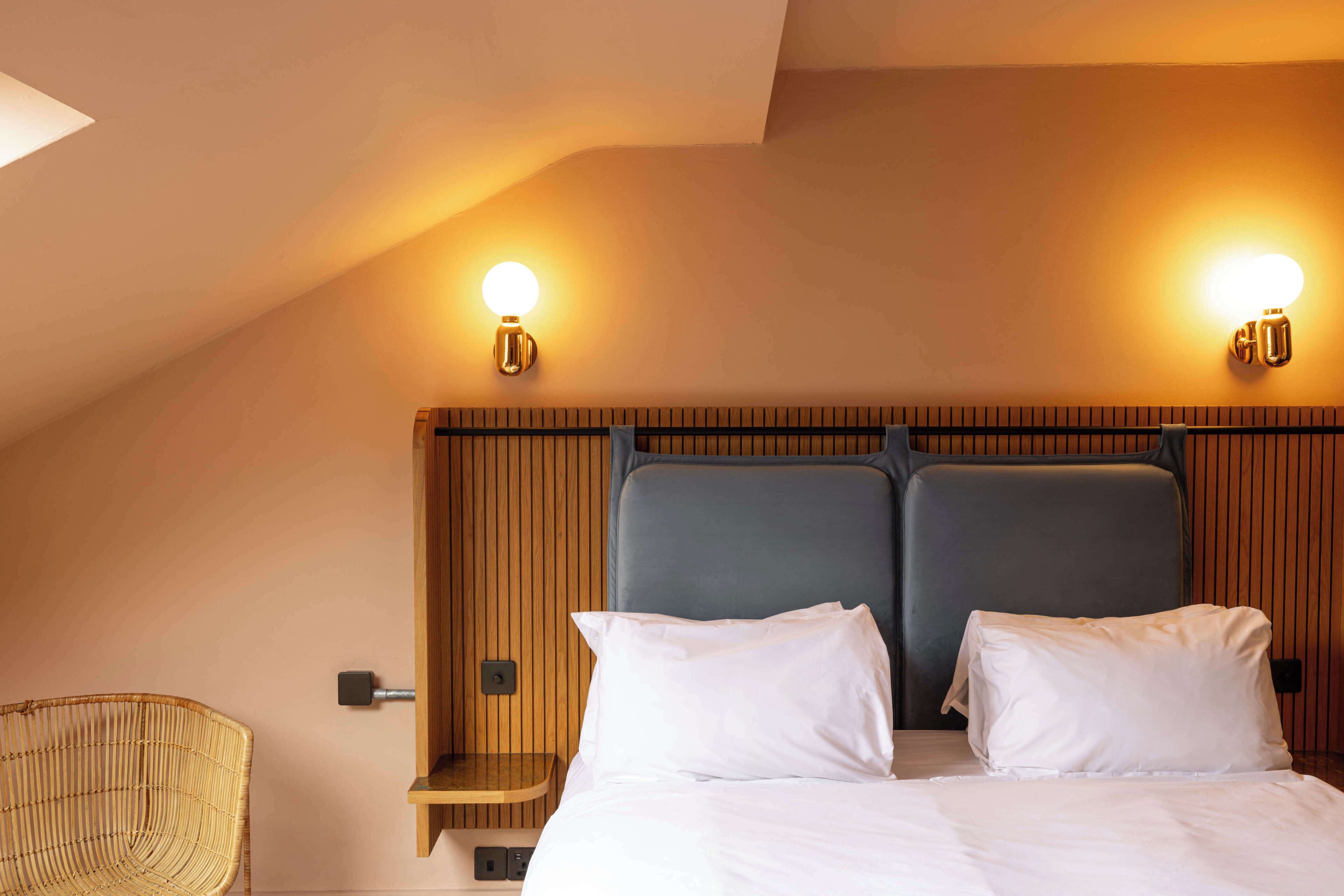 Habitación de hotel con cama, dos lámparas de pared, cabecera de madera y una silla de mimbre.