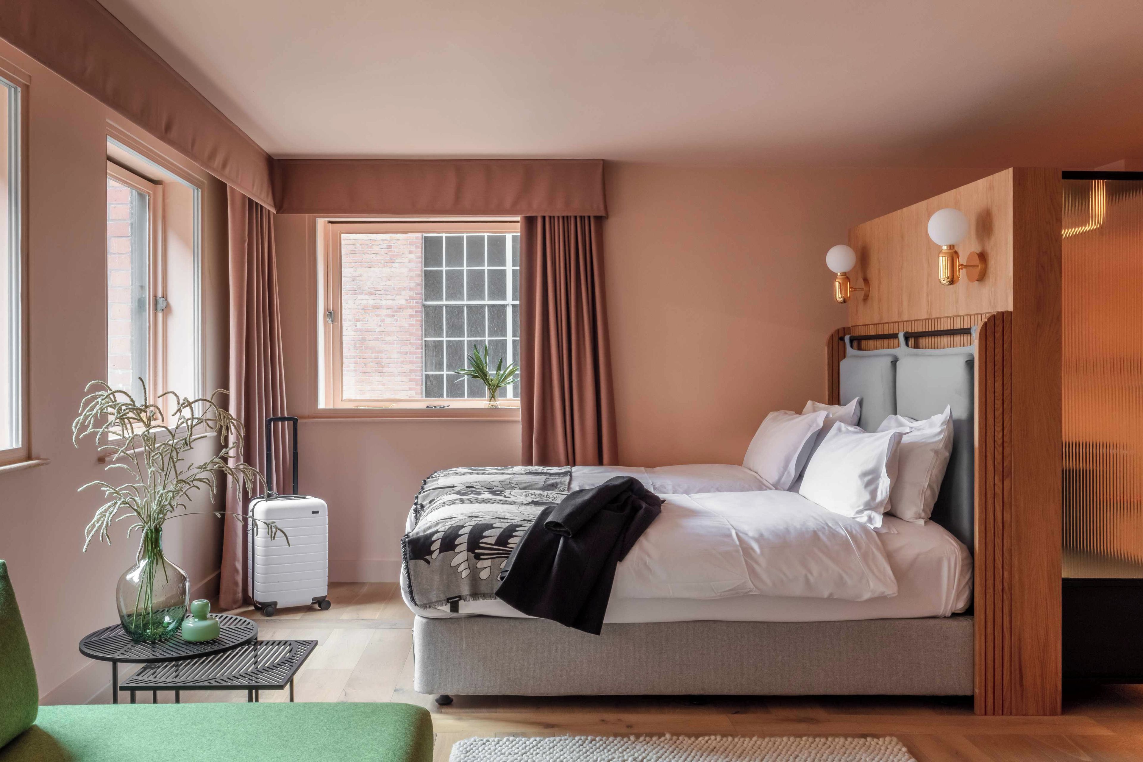 Una camera da letto con un letto, una valigia, una sedia verde, un tavolino con un vaso e una finestra con tende.