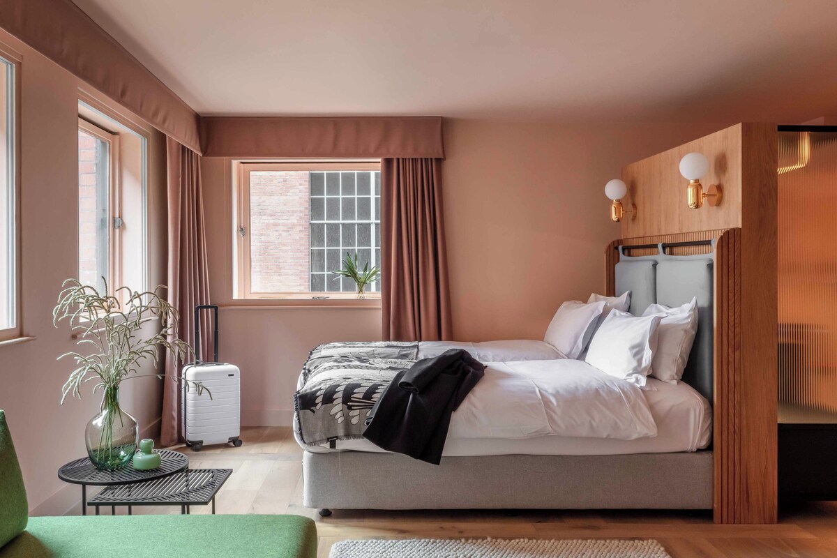 A bedroom with a bed, a suitcase, a green chair, a small table with a vase, and a window with curtains.