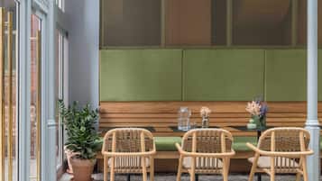 Een zithoek met een houten bank en drie rieten stoelen, een bijzettafeltje met een vaas en een karaf, en een plant in pot.