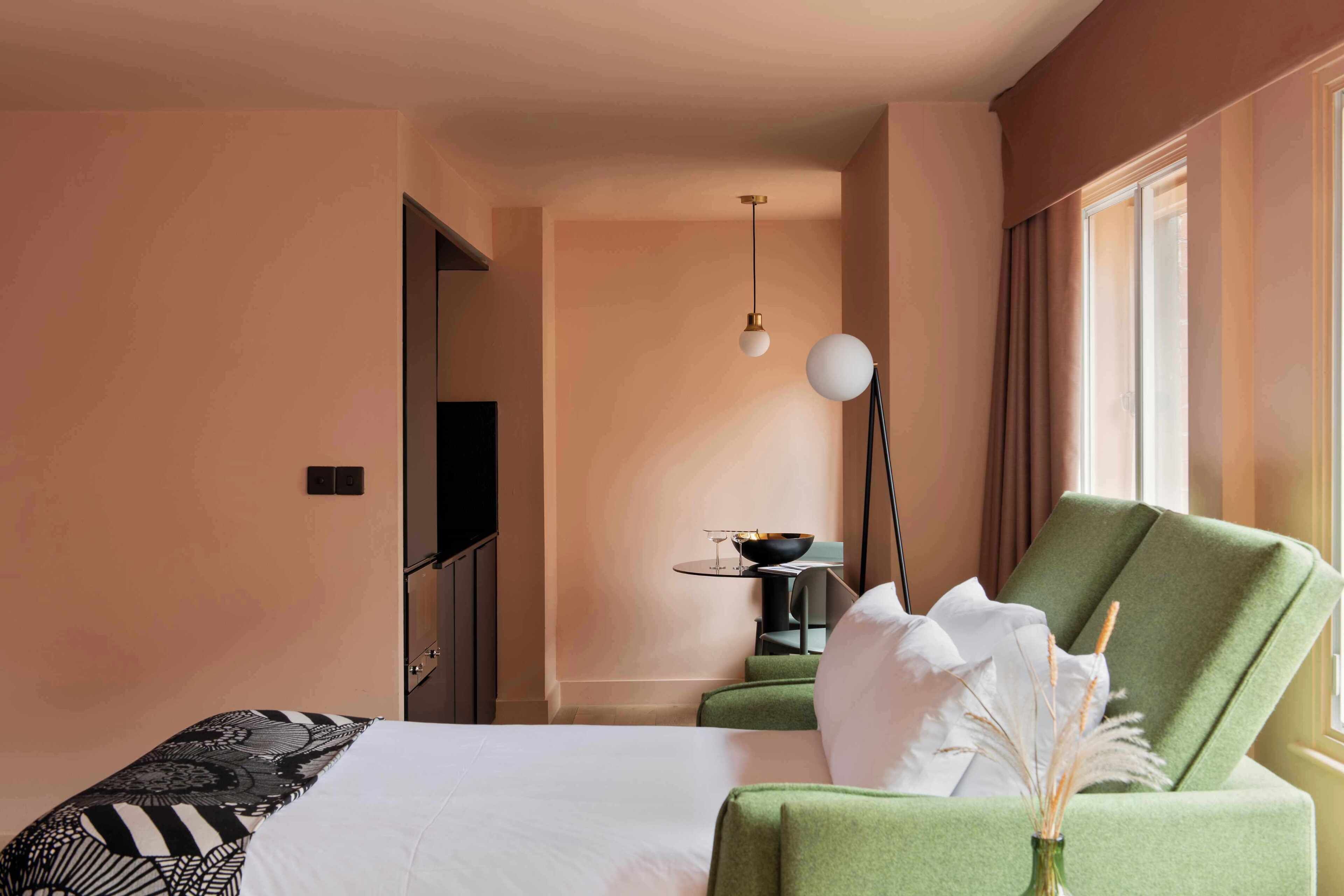 A hotel room with a bed, a green armchair, a small table with a bowl and glasses, and a hanging lamp.