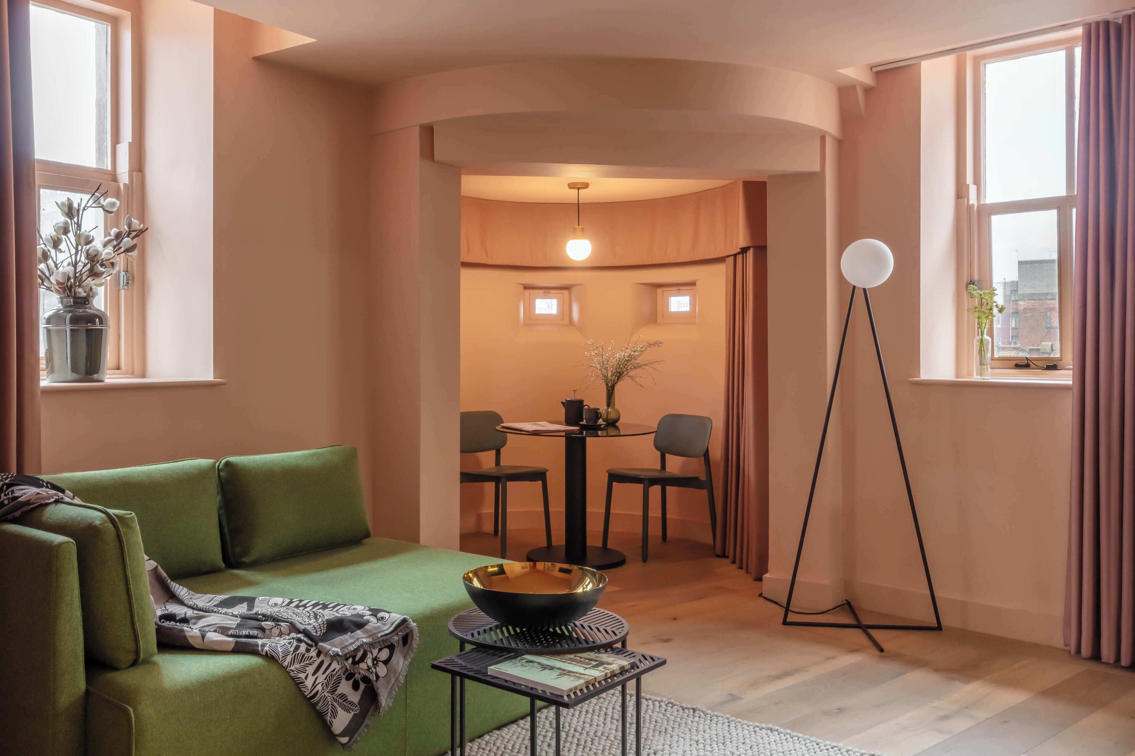 A modern living room with a green sofa, a small round table with a bowl, a floor lamp, and a dining area with chairs and a table.