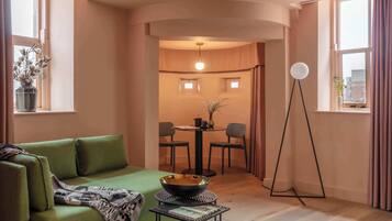 A modern living room with a green sofa, a small round table with a bowl, a floor lamp, and a dining area with chairs and a table.