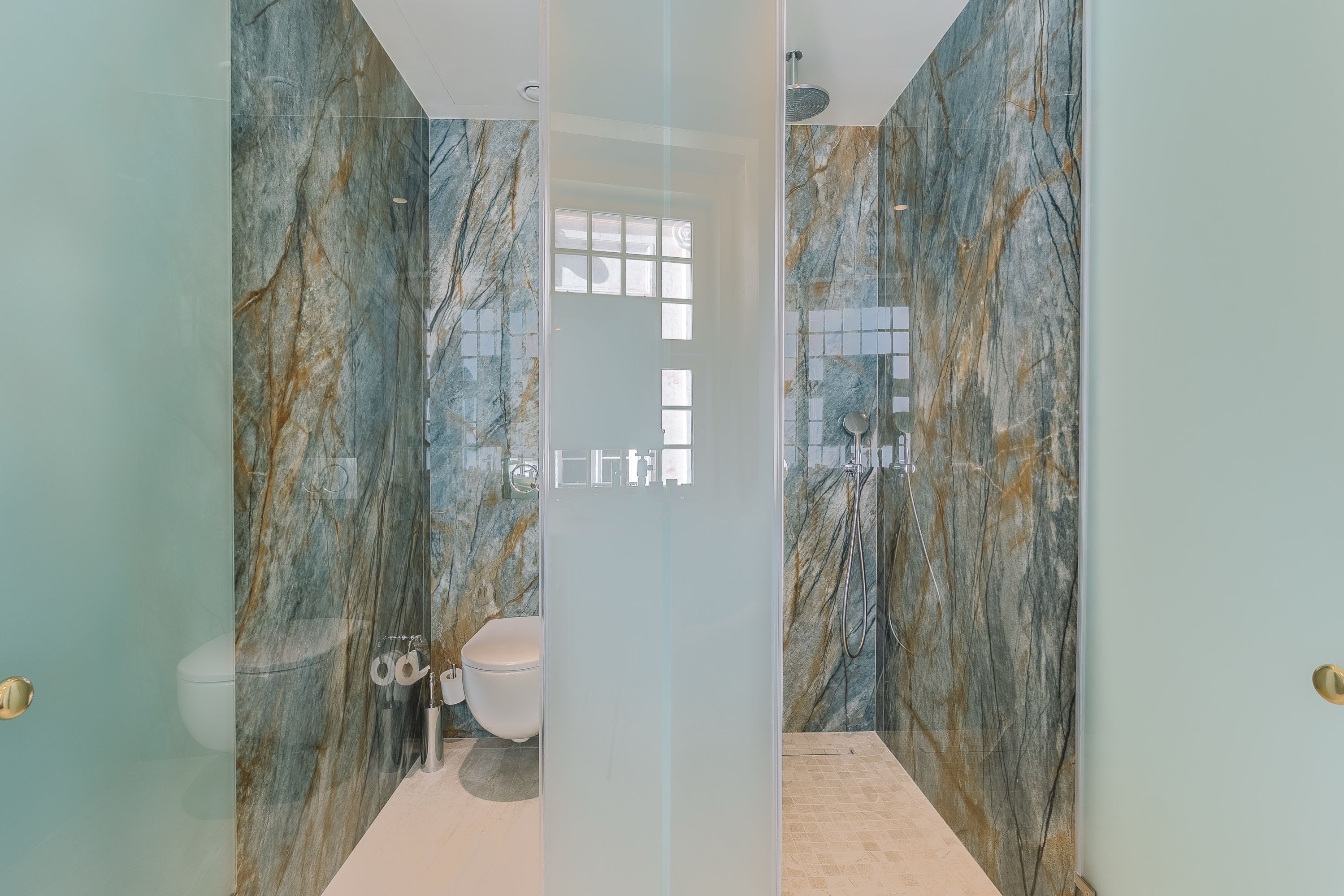 Un baño con puerta de vidrio, paredes de mármol, inodoro y zona de ducha.