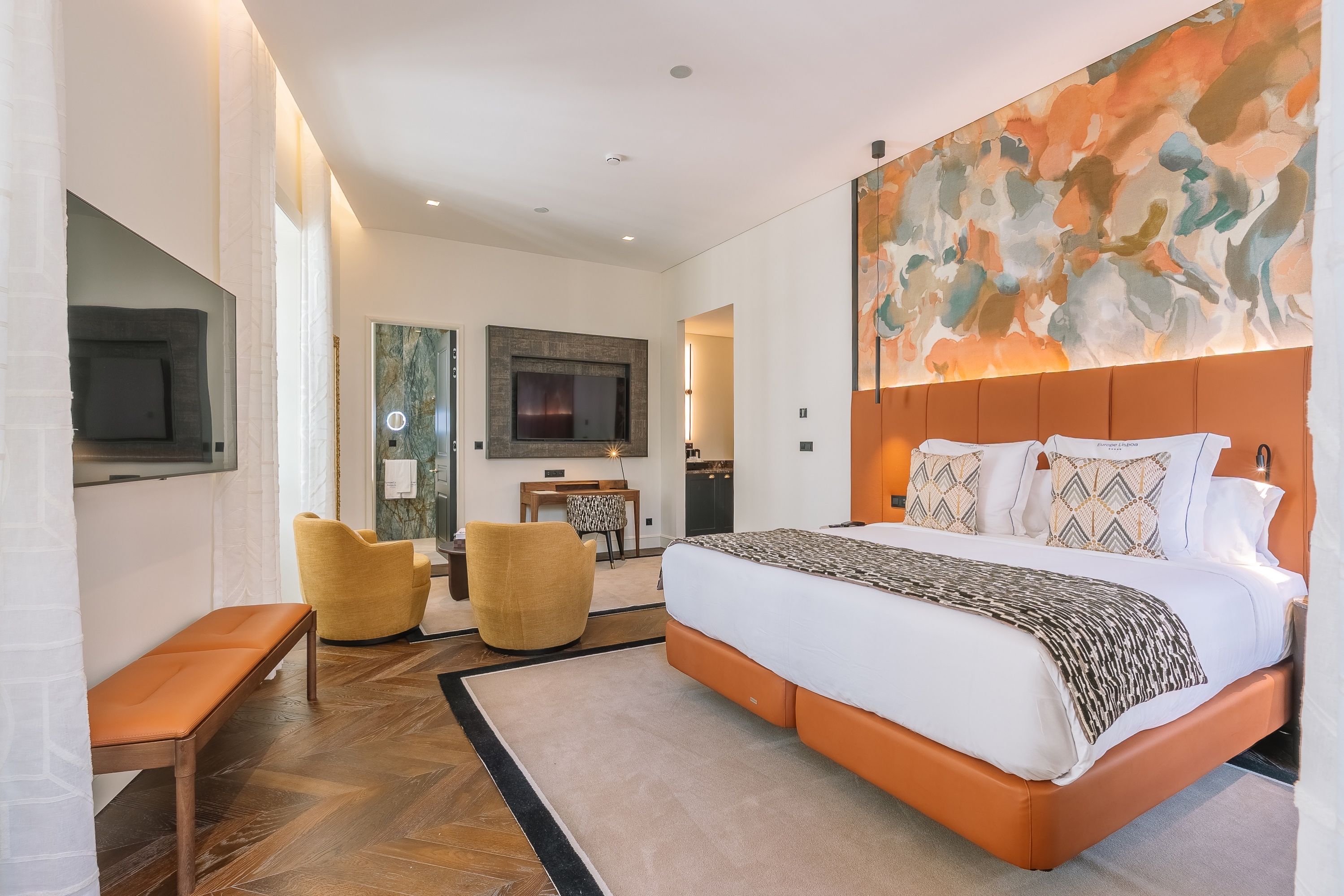 Een moderne hotelkamer met een groot bed, een flatscreen televisie, een zithoek met twee stoelen en een groot abstract schilderij aan de muur.
