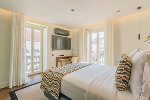 Habitación de hotel con cama, escritorio, silla, televisor y balcón con cortinas.
