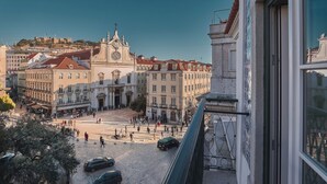 Uma praça histórica com uma igreja imponente, ruas de pedra e vista para uma colina com um castelo.
