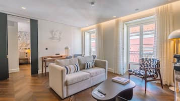 Een moderne woonkamer met een bank, fauteuil en salontafel, een eethoek met tafel en stoelen, en grote ramen met gordijnen.