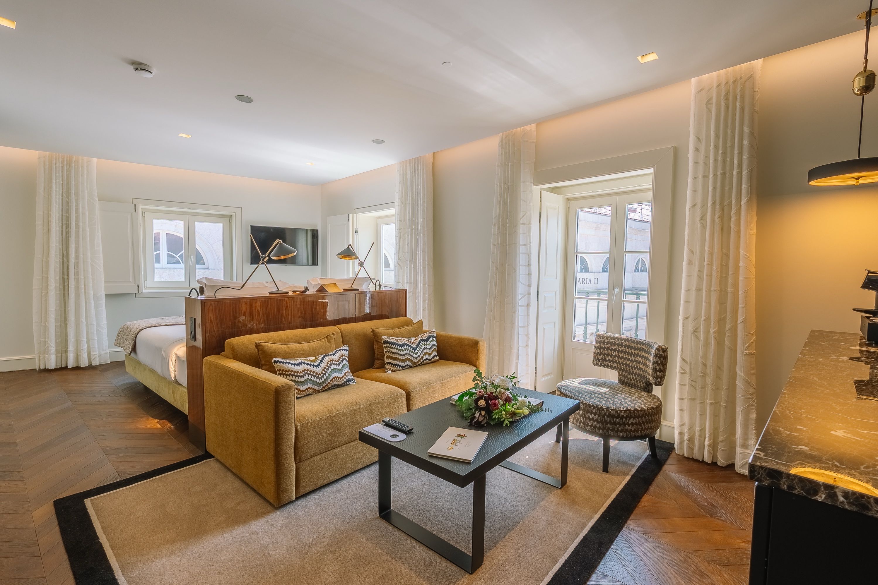 A modern hotel room with a sofa, a coffee table, a chair, a TV, and a window with curtains.