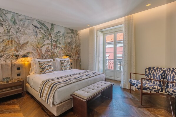 Habitación de hotel con cama, banqueta, silla, mesita de noche y pared con un mural tropical.