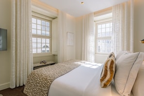 Habitación de hotel con una cama grande, dos ventanas con cortinas, un televisor de pantalla plana y una pequeña mesa con un ramo de flores.