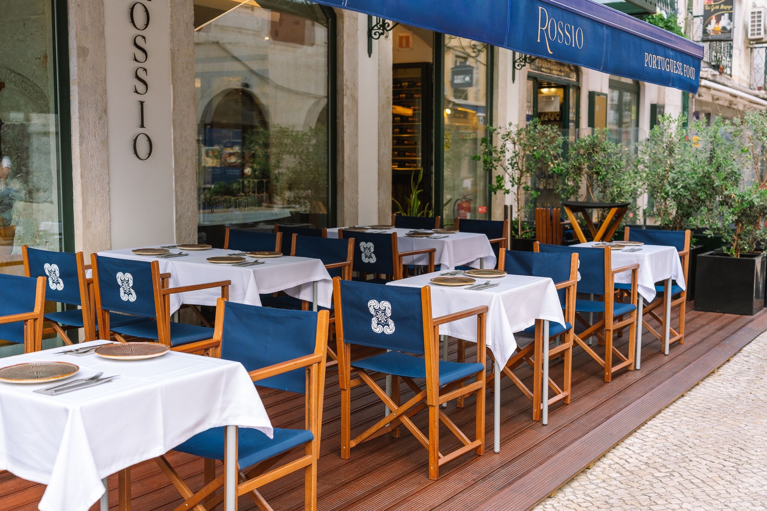 Un espacio de comedor al aire libre con mesas y sillas, un letrero que dice 'ROSSIO PORTUGUESE FOOD', y la fachada de un edificio con un letrero que dice 'ROSSIO'.