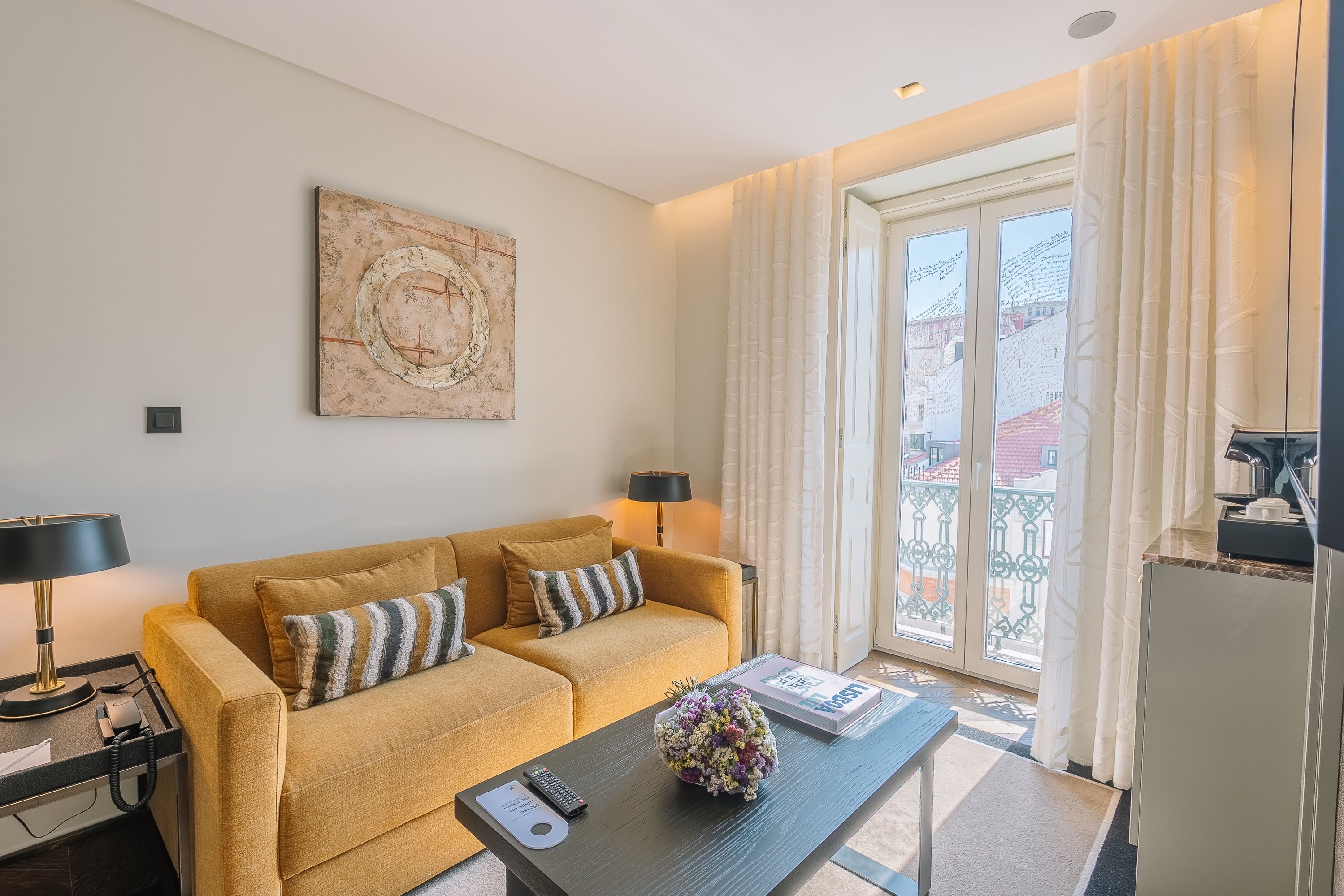 A living room with a sofa, two lamps, a coffee table with a remote and a book, and a large window with curtains.