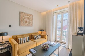 A living room with a sofa, two lamps, a coffee table with a remote and a book, and a large window with curtains. - Europe Lisboa Boutique Hotel (Lisbon)