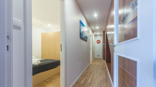 A hotel corridor with wooden flooring, a bed with a dark bedspread, a white door with a red "no entry" sign, and a wall-mounted painting.