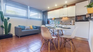 A modern kitchen with white cabinets, a dining table with chairs, and a grey sofa with blue cushions.