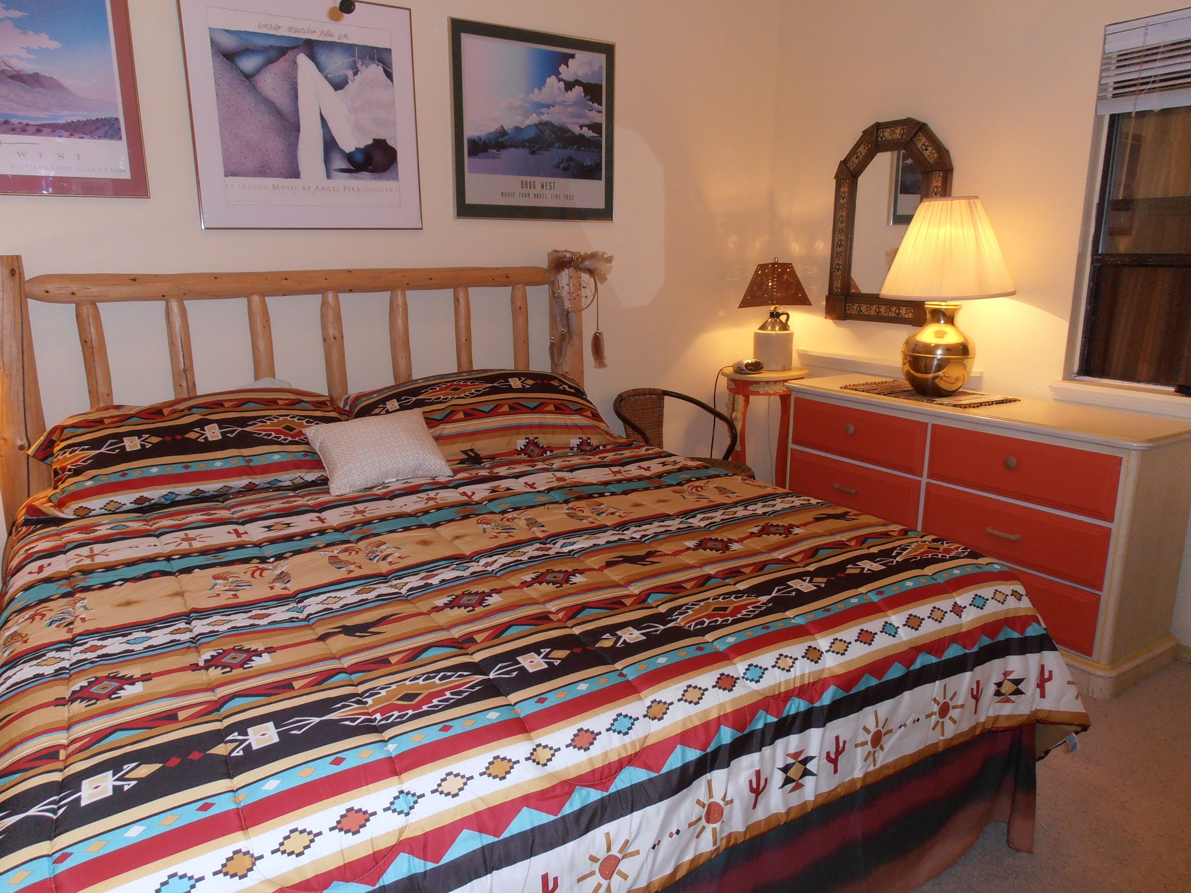 A bed with a patterned bedspread, a wooden headboard, a nightstand with a lamp, and a dresser with drawers.