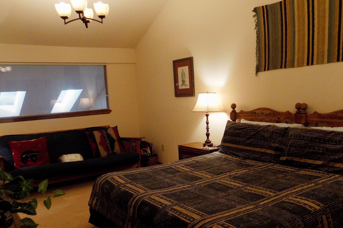 A bedroom with a large bed, a nightstand with a lamp, a TV, a window with blinds, and a wall tapestry.