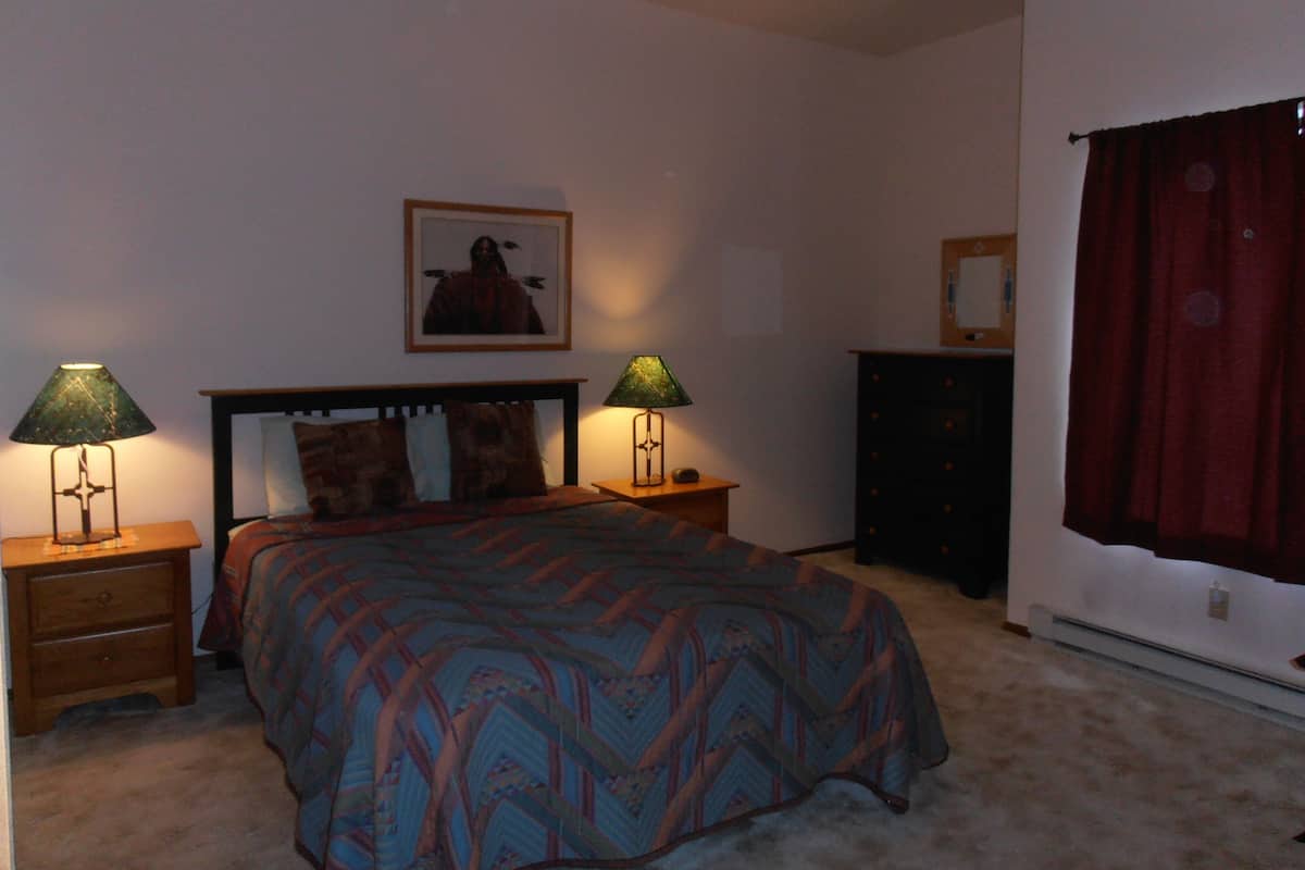 A bedroom with a bed, two bedside tables with lamps, a dresser, a framed picture on the wall, and a window with curtains.