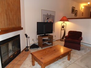 A living room with a television on a wooden stand, a red armchair, a fireplace, and a floor lamp. - Angel Fire Chalet (Angel Fire)