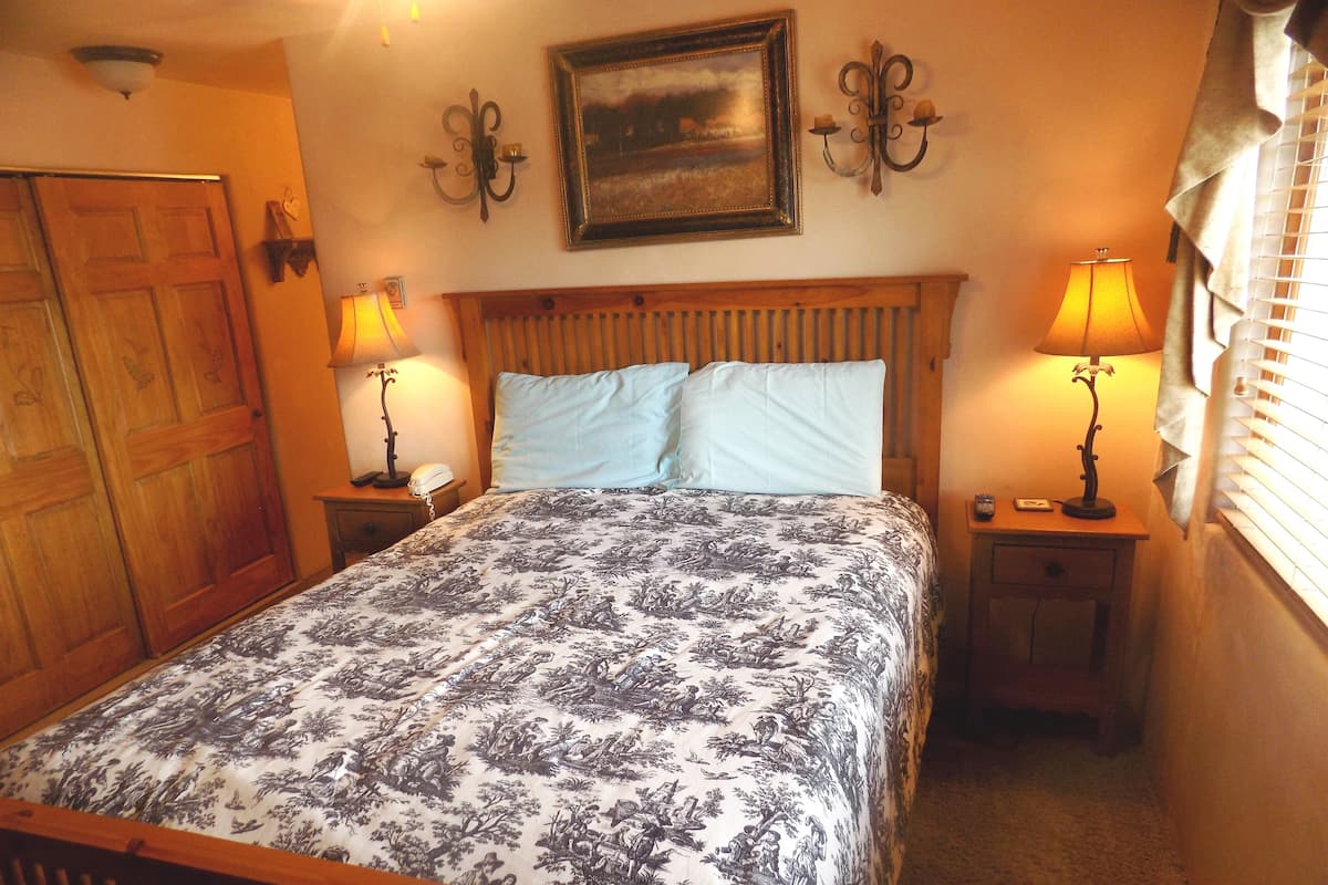 A bedroom with a bed, two bedside tables, a nightstand, a wooden headboard, a painting, wall sconces, and a window with blinds.