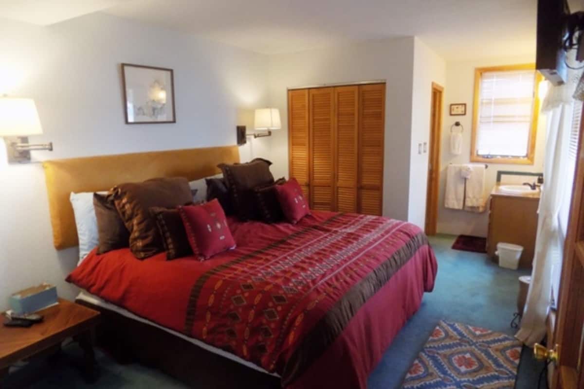 A neatly arranged bedroom with a bed, bedside lamps, a wardrobe, and a bathroom visible through an open door.