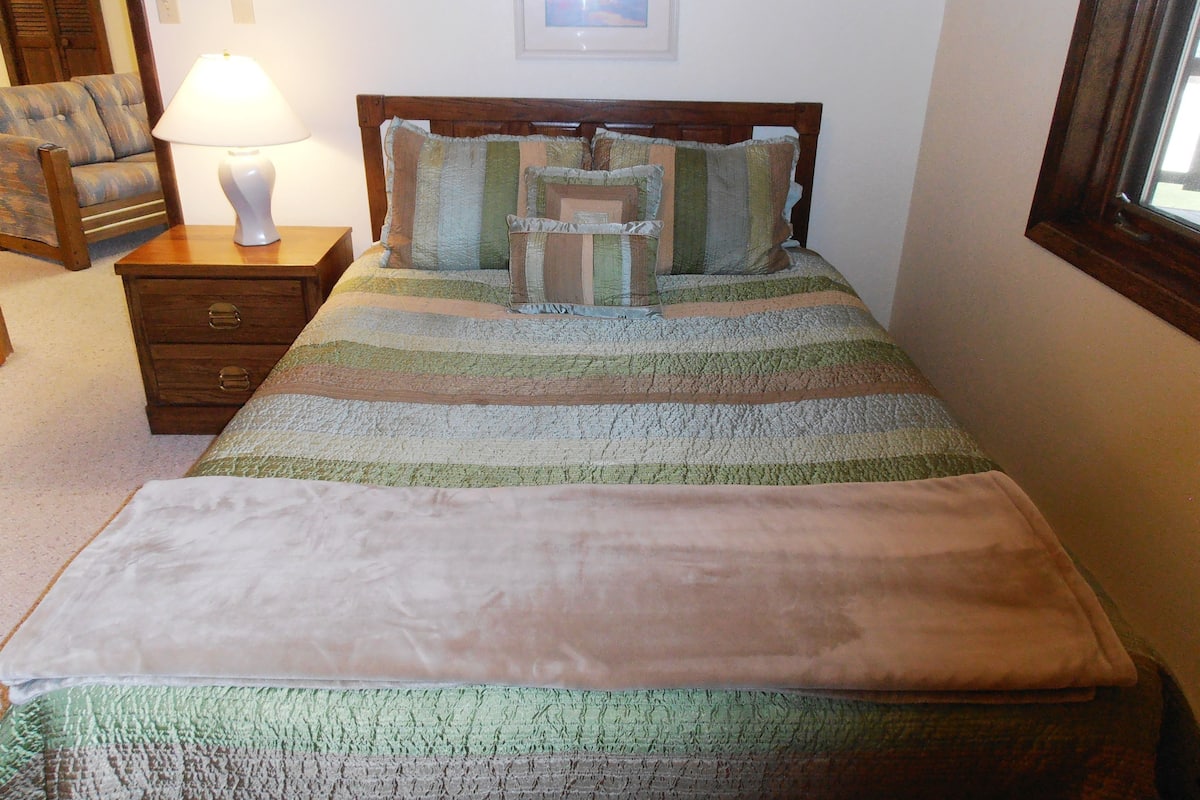 A bedroom with a bed, a nightstand, a lamp, a chair, and a window.