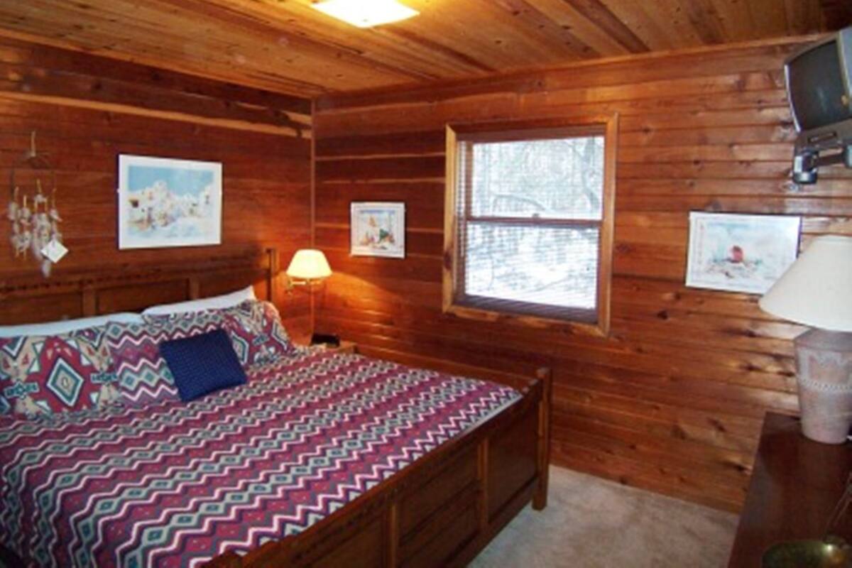 A wooden cabin room with a bed, a window, and a television on a wooden stand.