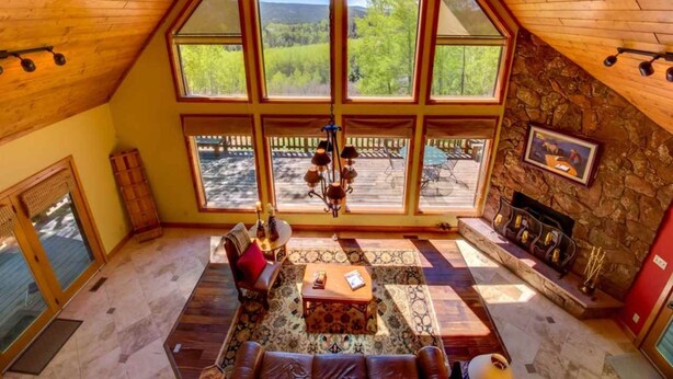 A spacious loft with a high ceiling, large windows offering a view of a forested area, a fireplace, and a cozy seating area. - Country Club Homes (Angel Fire)