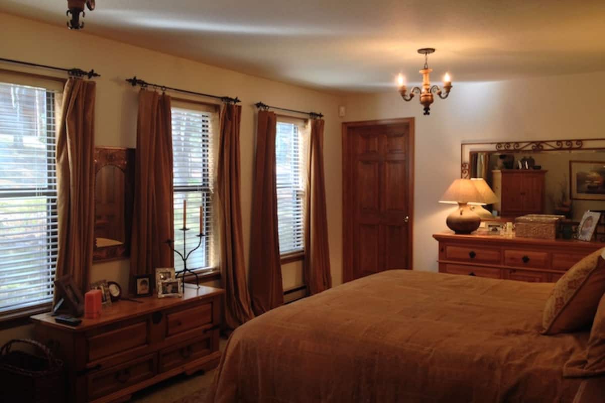 A bedroom with a large bed, wooden dresser, and a nightstand with a lamp. There are windows with curtains and a door in the background.