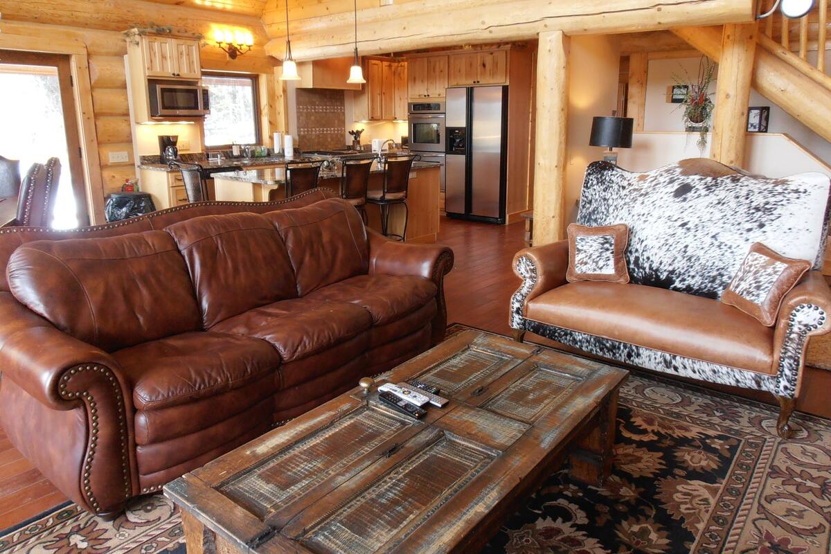 A cozy log cabin interior with a brown leather sofa, a patterned coffee table, and a rustic dining area.