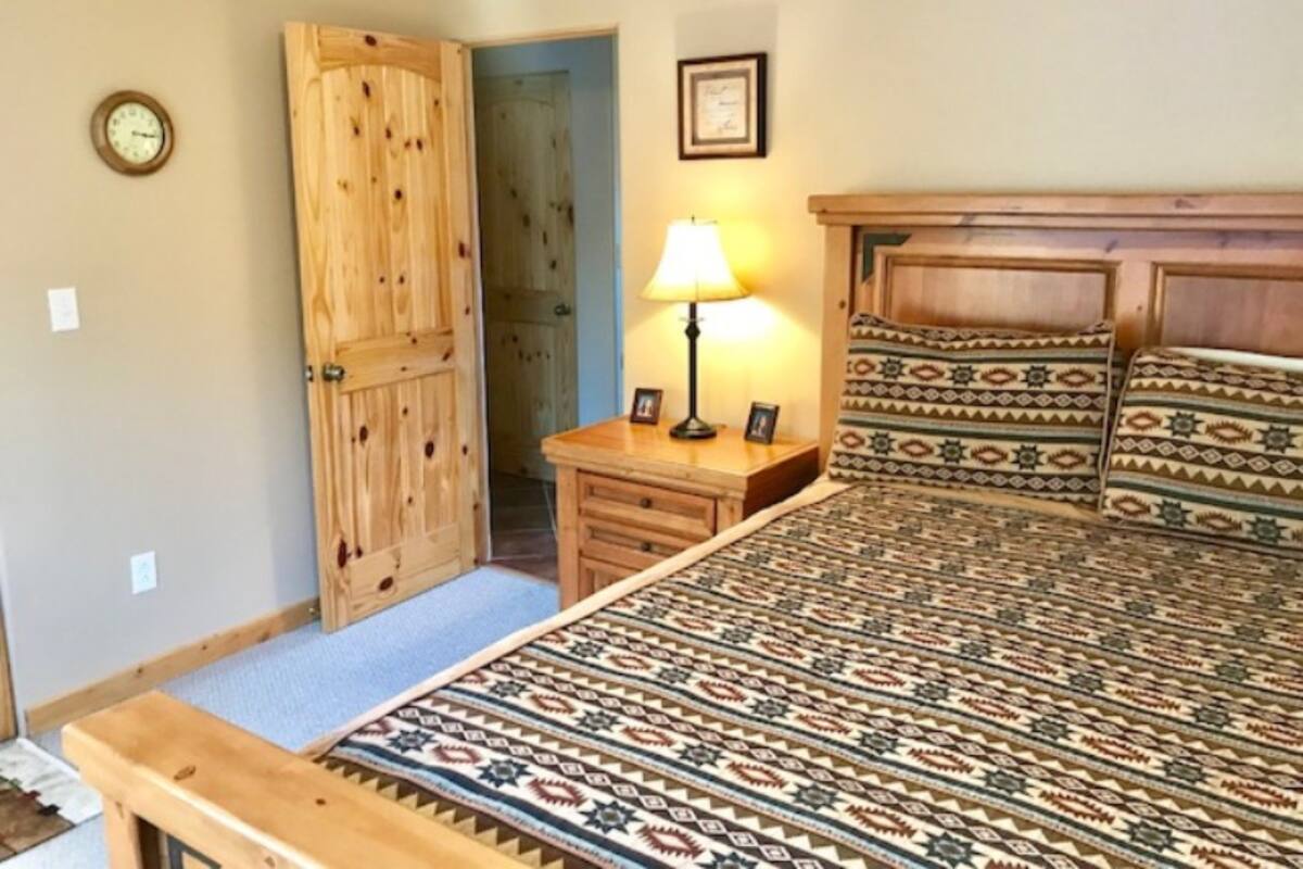 A bedroom with a wooden bed, a bedside table with a lamp, a clock on the wall, and a door leading to another room.