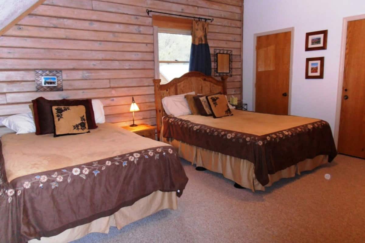 A bedroom with two beds, wooden walls, a window with curtains, a door, and framed pictures on the wall.
