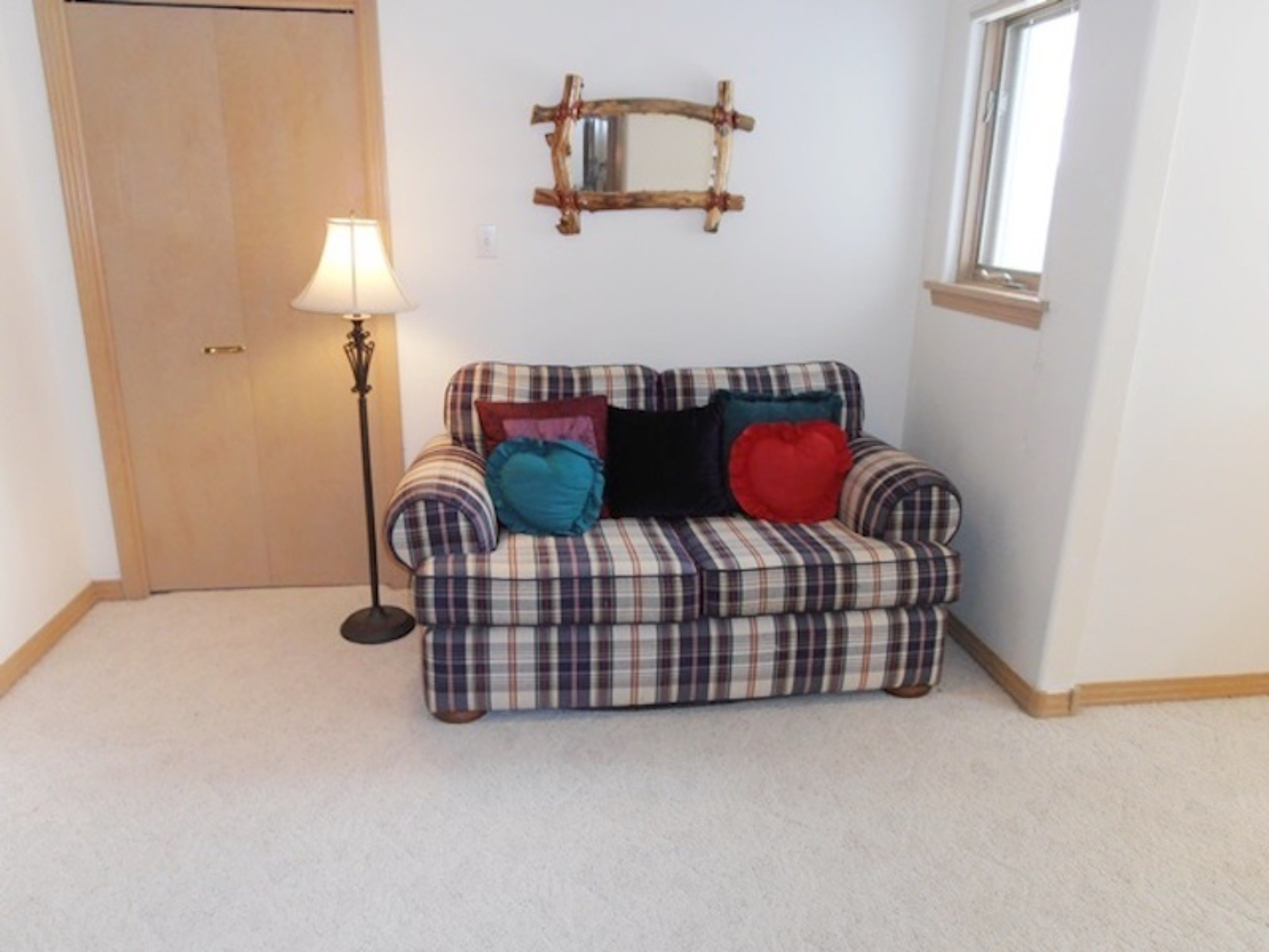 A plaid sofa with multiple cushions, a floor lamp, a wooden door, and a mirror on the wall.