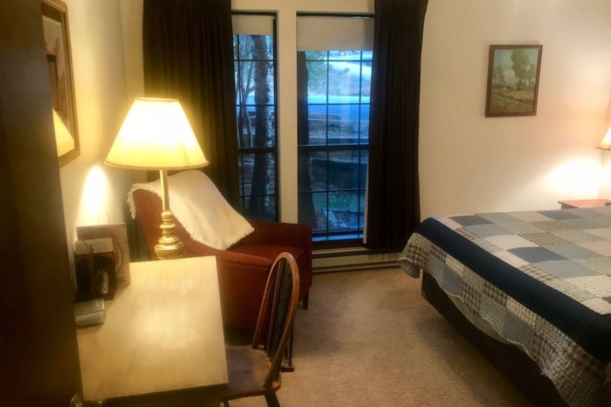 A hotel room with a bed, a desk with a lamp, a chair, and a window with curtains.