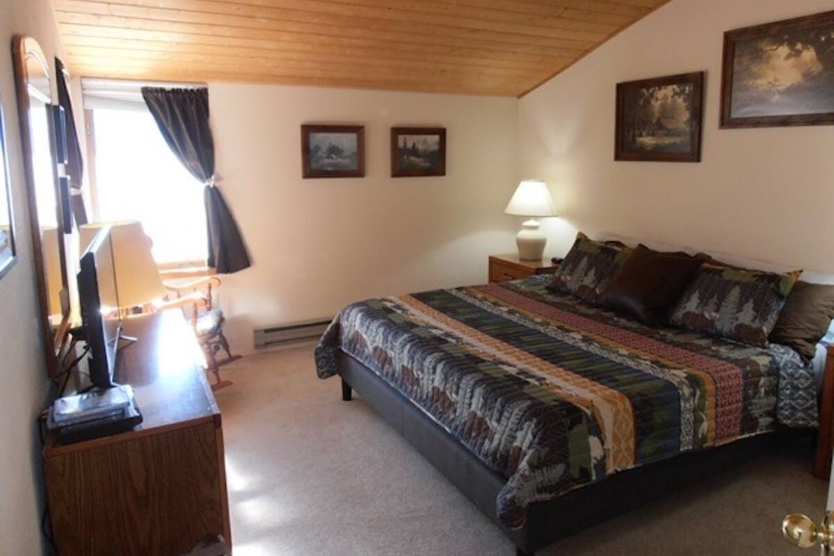 A bedroom with a wooden ceiling, a bed with a patterned bedspread, a nightstand with a lamp, and two framed pictures on the wall.