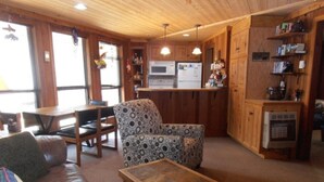 Een knus interieur van een chalet met een houten plafond, een eethoek, een gestreept fauteuil, een leren bank en een keuken met houten kastjes en een inbouwkoelkast.