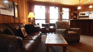 Een gezellige woonkamer met een leren bank, een fauteuil met een patroon, en een houten salontafel. Ook is een lamp en een eettafel met stoelen zichtbaar.