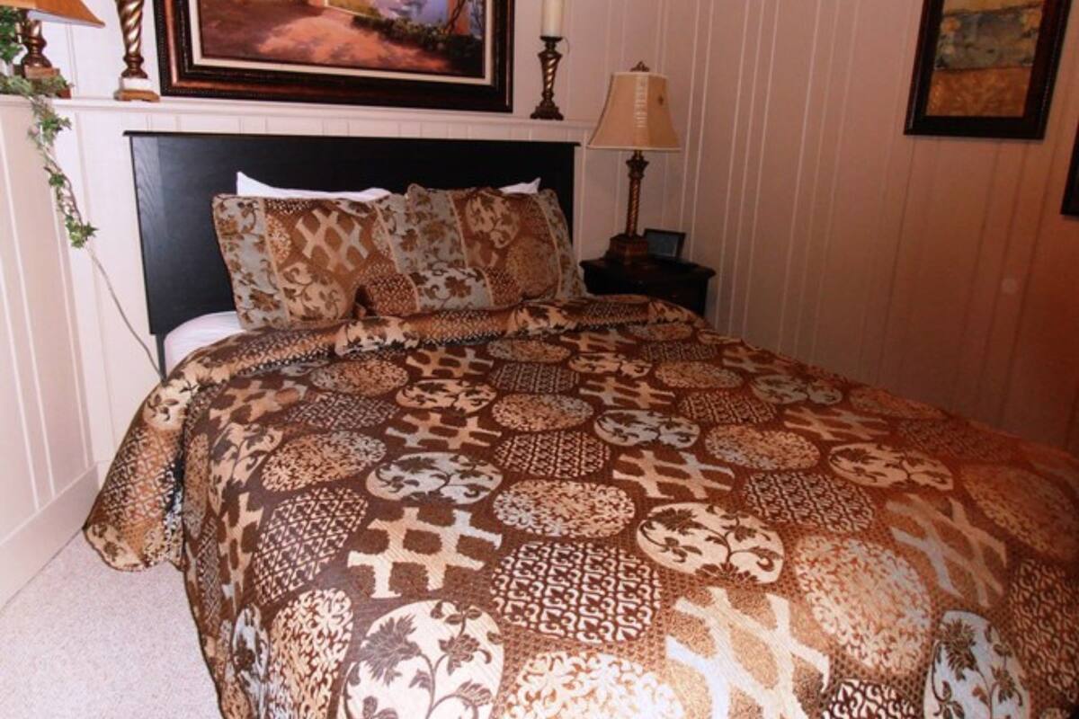 A bedroom with a patterned bedspread, a framed landscape painting above the bed, and a bedside table with a lamp.