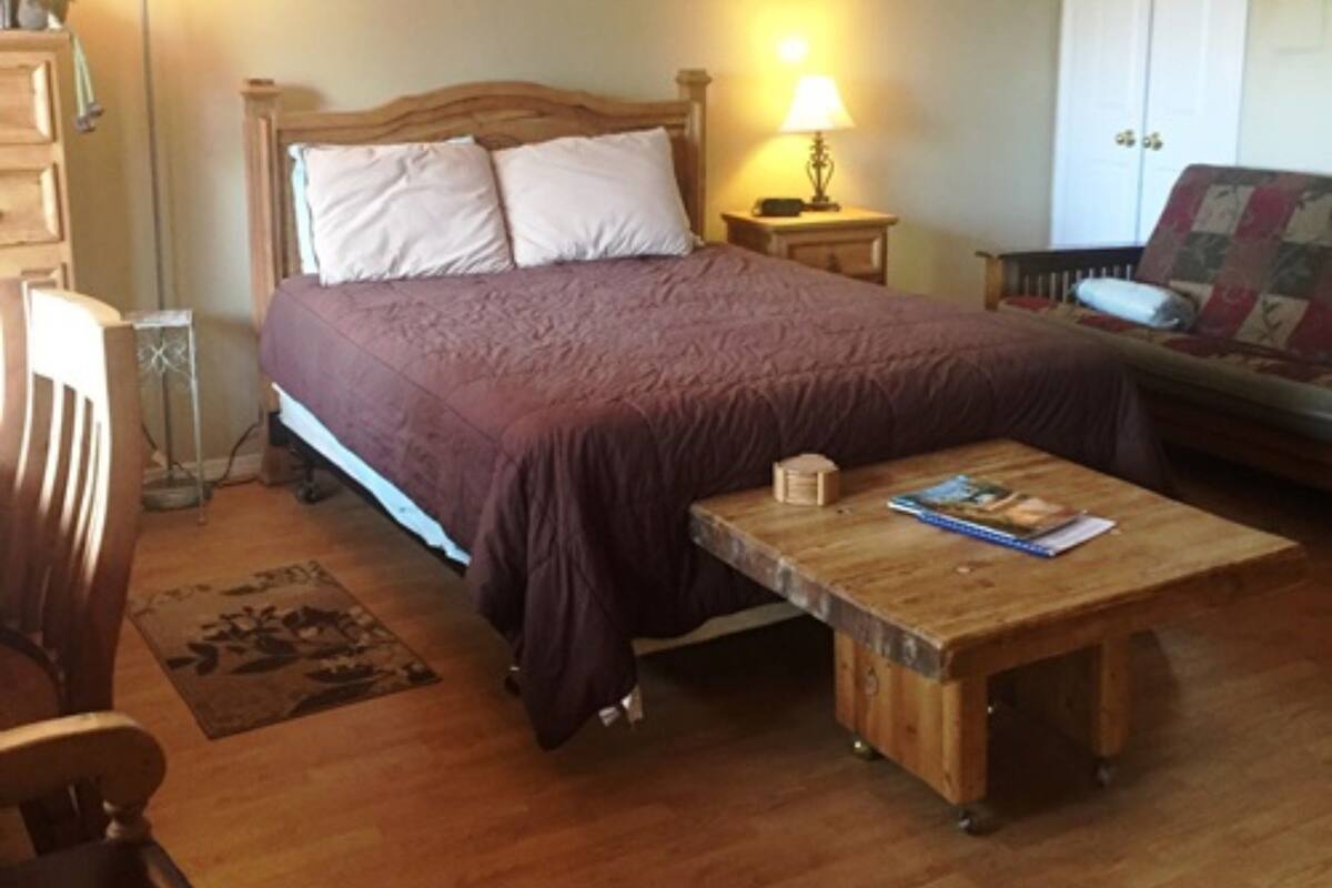 A bedroom with a wooden bed, a nightstand with a lamp, a wooden coffee table, a chair, and a wardrobe.