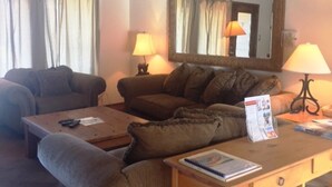 A living room with a sofa, a coffee table, and a desk with a lamp. There is a mirror on the wall and a window with curtains. - Ski Run (Angel Fire)